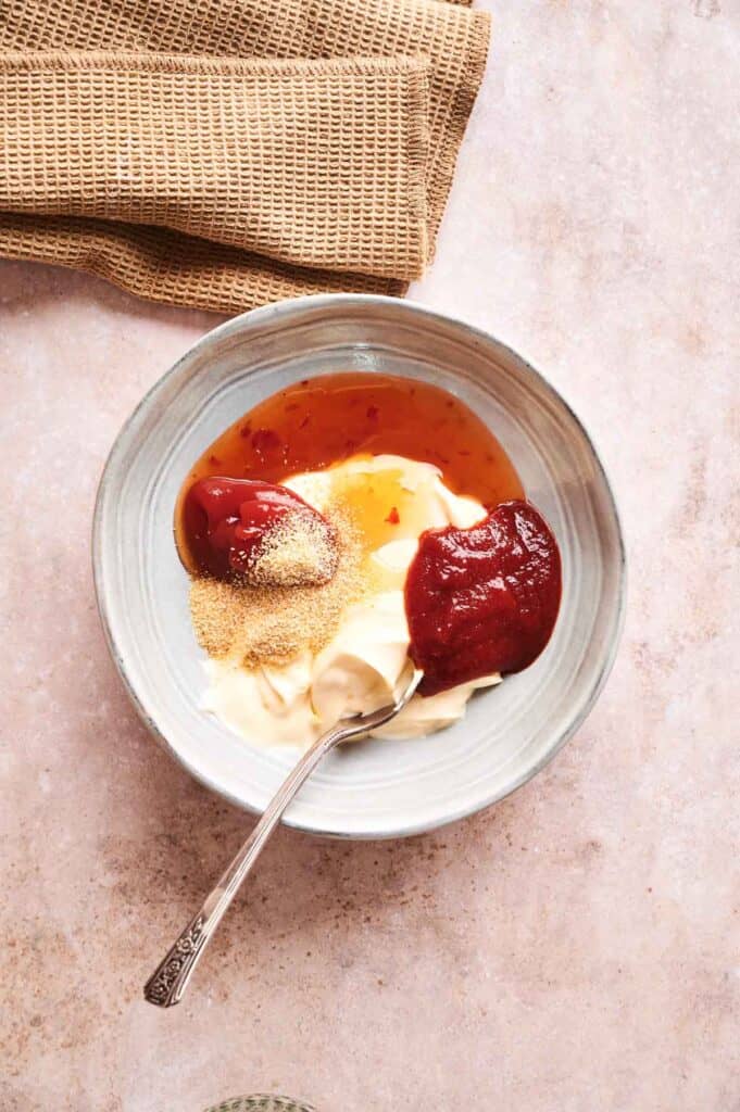 A bowl containing Boom Boom Sauce&mdash;a mix of mayonnaise, ketchup, sweet chili sauce, tomato paste, and granulated seasoning&mdash;with a spoon inside. A beige cloth sits nearby on the countertop.