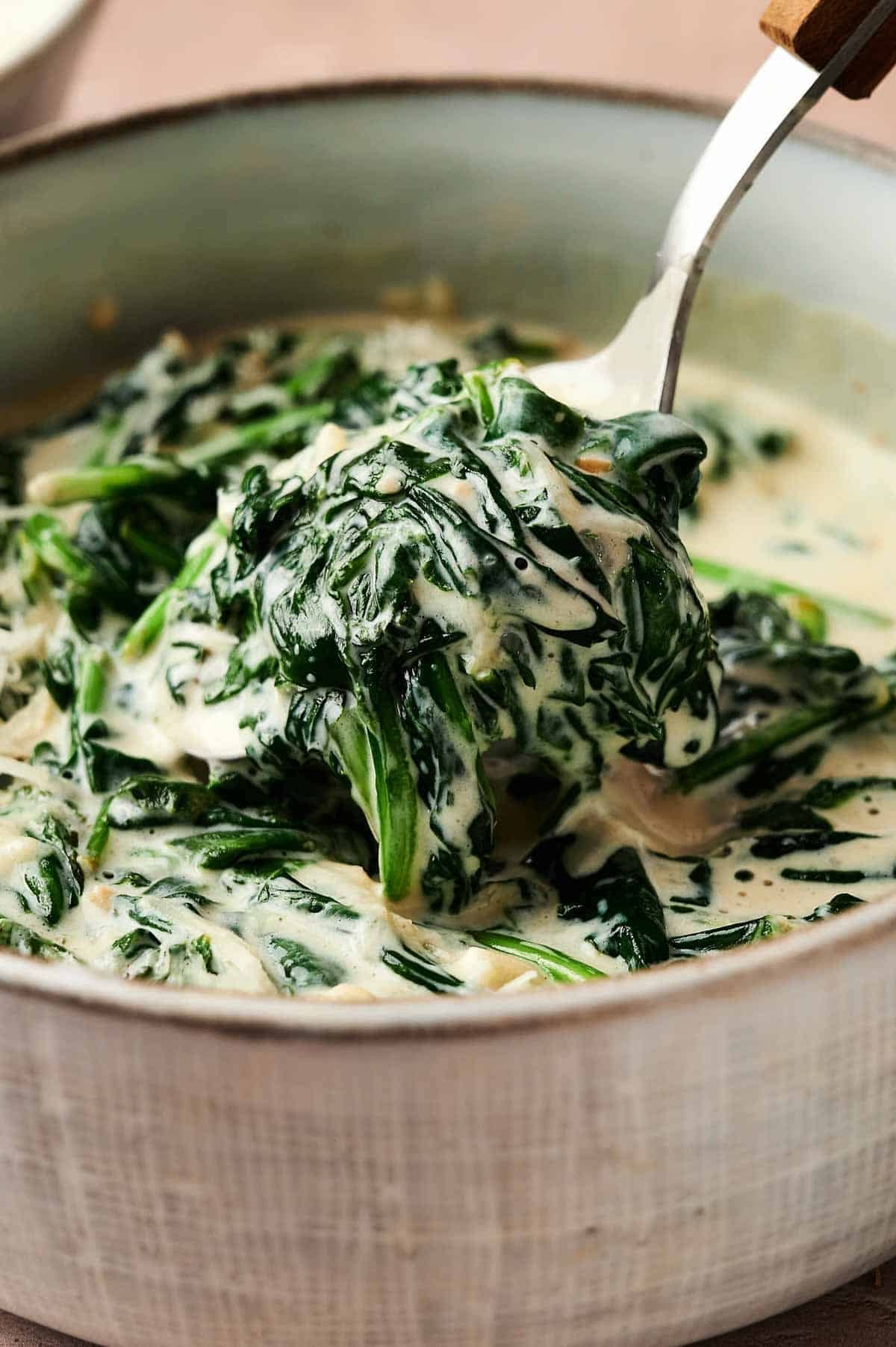 Creamed Spinach being lifted from a pot with a spoon, showing tender wilted spinach leaves enveloped in a rich, creamy sauce.