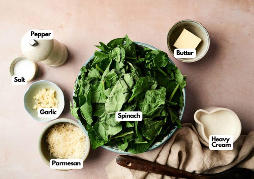 A bowl of fresh spinach, perfect for making Creamed Spinach, is surrounded by bowls of butter, heavy cream, grated Parmesan, minced garlic, salt, and a pepper grinder on a light surface.