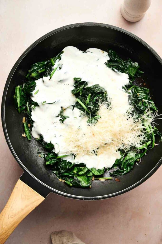 A frying pan with creamed spinach, a creamy white sauce, and grated cheese on top rests on a light surface.