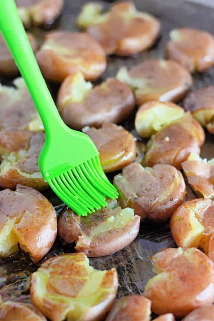 A green silicone brush spreads oil over smashed, cooked potatoes on a baking sheet, prepping them for Crispy Garlic Parmesan Smashed Potatoes.