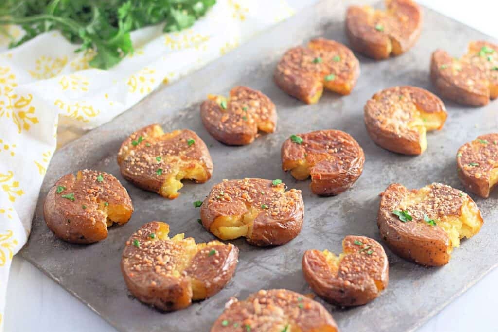 A baking sheet with several Crispy Garlic Parmesan Smashed Potatoes, topped with herbs and seasoning, arranged in rows.