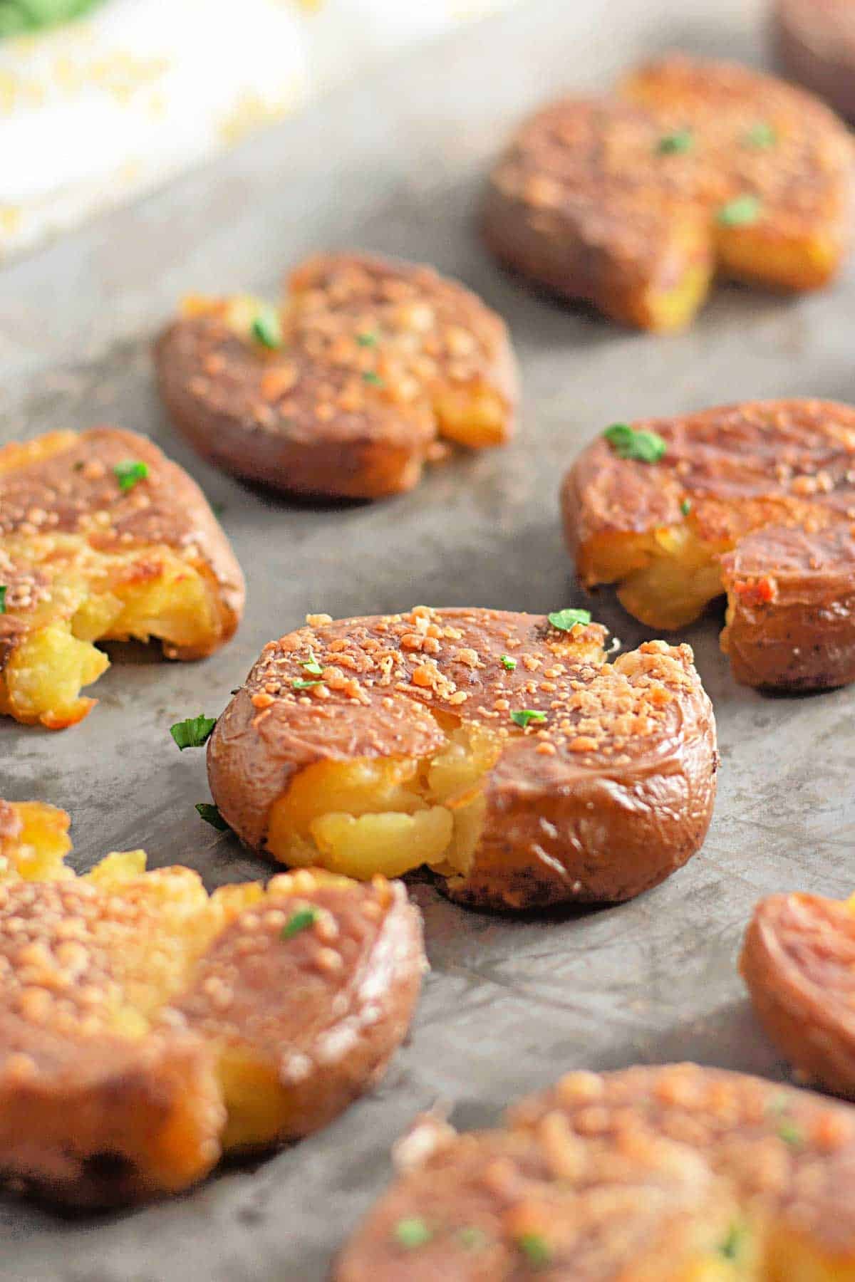 Several Crispy Garlic Parmesan Smashed Potatoes are arranged on a baking sheet, their golden edges topped with seasoning and garnished with chopped herbs.