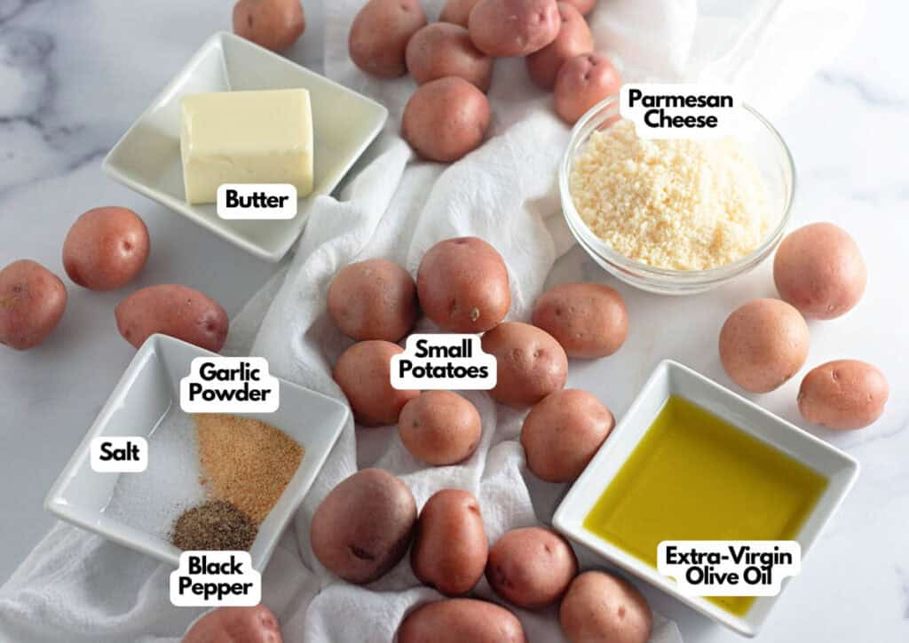 Ingredients for Crispy Garlic Parmesan Smashed Potatoes—small red potatoes, butter, parmesan cheese, extra-virgin olive oil, garlic powder, salt, and black pepper—are arranged separately on a white surface.