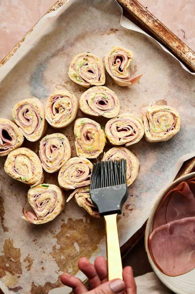Several Ham and Cheese Pinwheels are arranged on a parchment-lined baking sheet, as a hand with a pastry brush prepares to glaze them. A bowl of sliced ham sits nearby, ready for the next batch.
