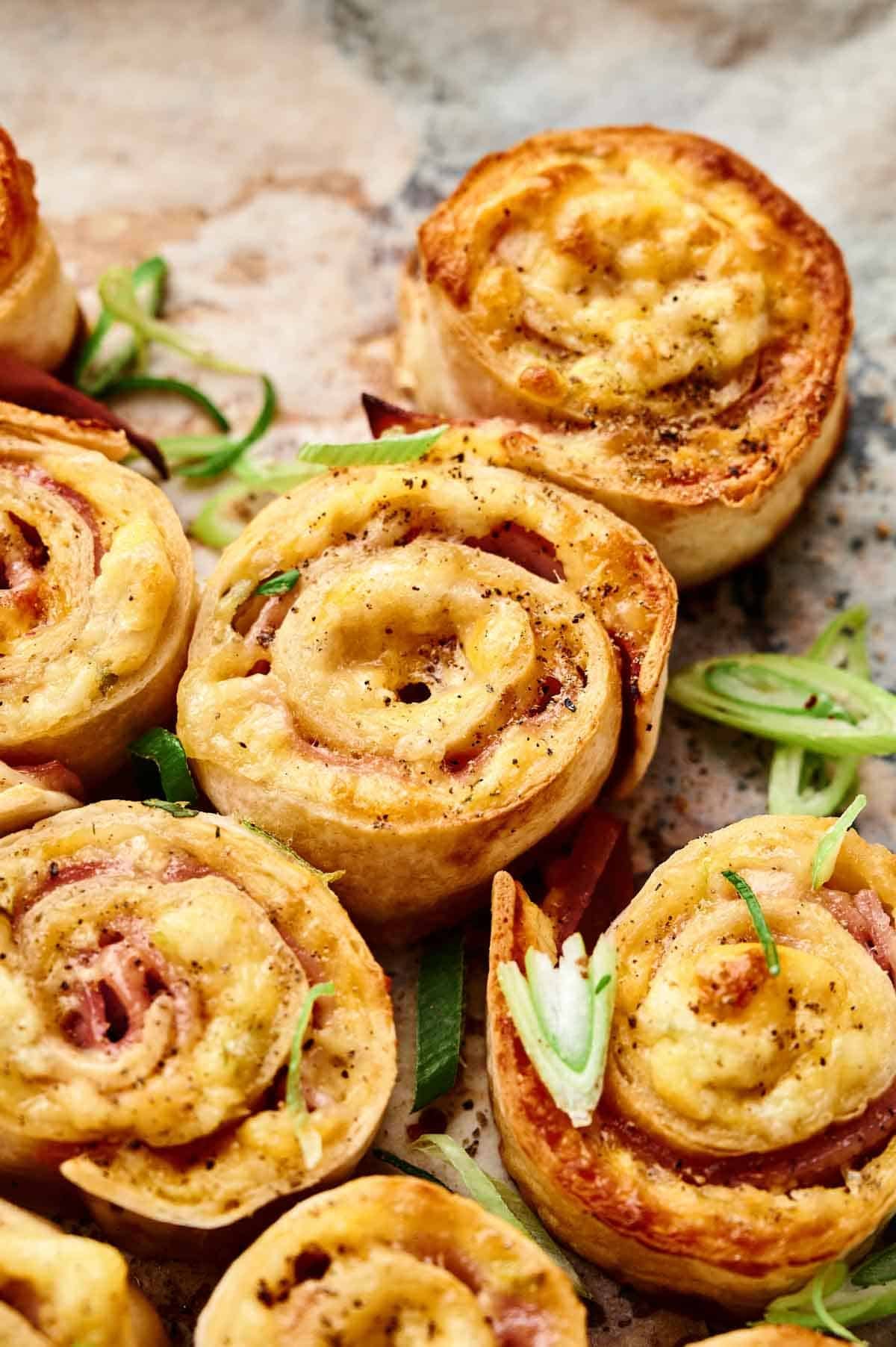 Close-up of Ham and Cheese Pinwheels made with pastry, melted cheese, and ham, garnished with sliced green onions on a parchment-lined surface.