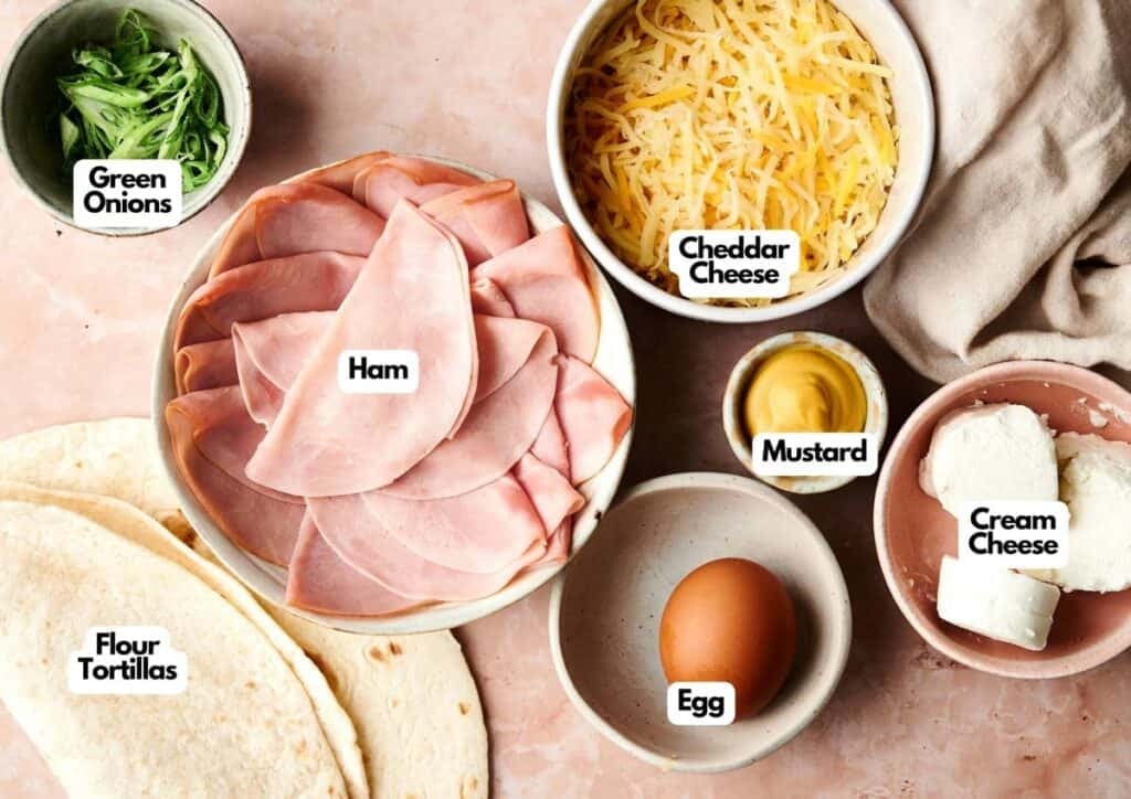 Bowls containing ingredients for Ham and Cheese Pinwheels&mdash;green onions, cheddar cheese, ham, mustard, cream cheese, an egg, and flour tortillas&mdash;are arranged on a light surface.