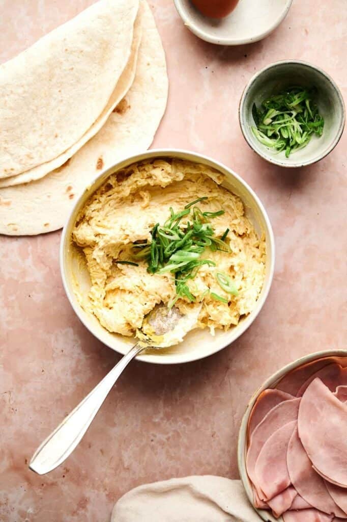 Bowl of scrambled eggs topped with sliced green onions, surrounded by tortillas, a bowl of chopped green onions, sliced ham on a plate, and Ham and Cheese Pinwheels for a delicious breakfast spread.
