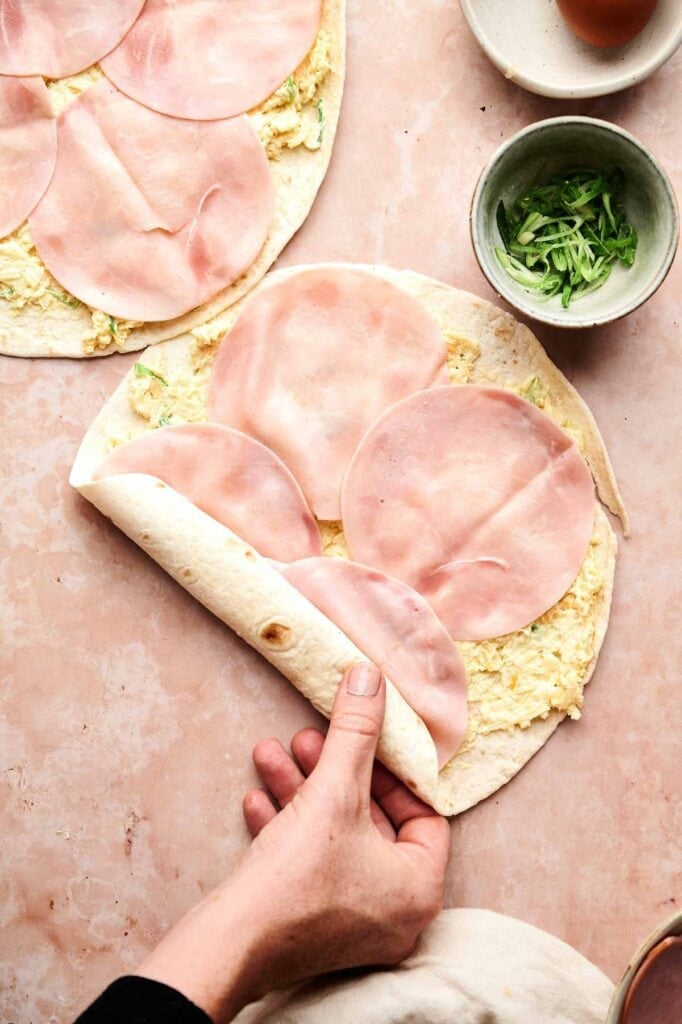 A hand prepares Ham and Cheese Pinwheels by rolling a tortilla topped with shredded cheese mixture and slices of ham; bowls of green onions and an egg sit nearby.
