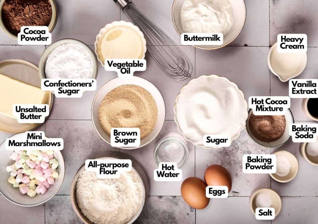 Various baking ingredients in bowls, including flour, sugars, cocoa powder, eggs, butter, buttermilk, marshmallows, and heavy cream&mdash;everything you need to make Hot Chocolate Cupcakes&mdash;are labeled with text on a tiled surface.