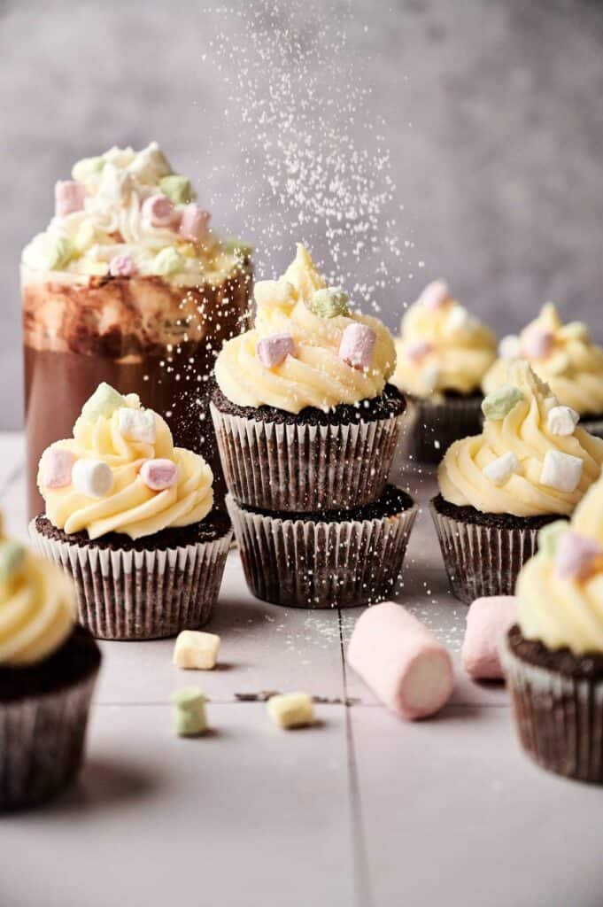 Hot Chocolate Cupcakes with yellow frosting and mini marshmallows are arranged on a surface, as powdered sugar is being sprinkled over them.