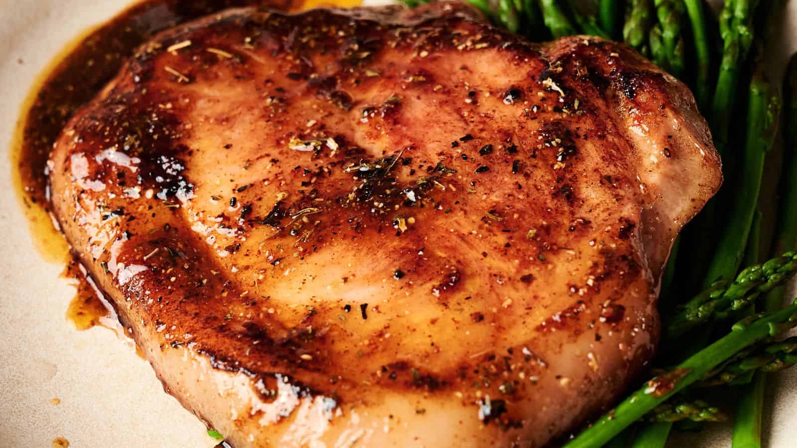 Close-up of a seasoned, cooked pork chop with a caramelized crust, served on a plate with steamed asparagus spears.
