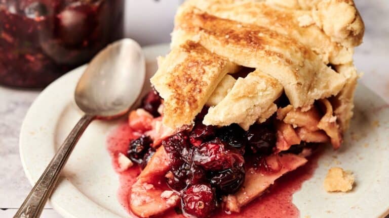 A slice of mixed berry pie with a flaky crust sits on a plate next to a spoon, with some filling spilling out.