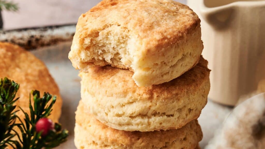 Three golden brown biscuits are stacked on a plate, with the top biscuit missing a bite. A small green decoration with red berries is visible in the foreground.