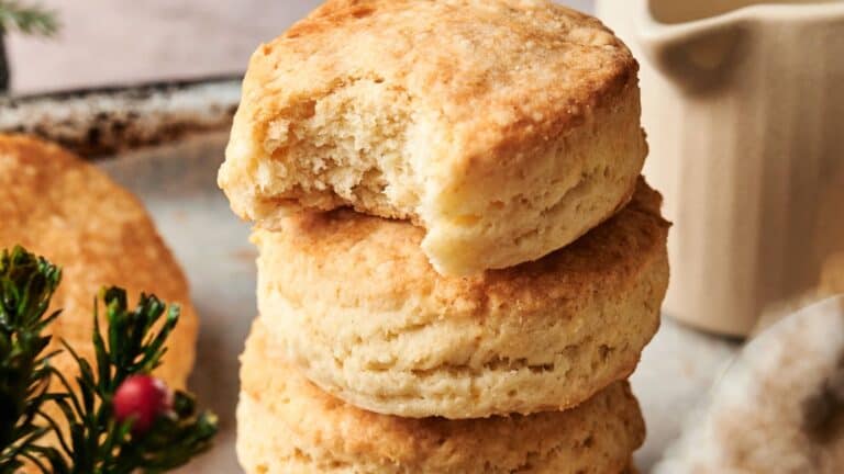 Three golden brown biscuits are stacked on a plate, with the top biscuit missing a bite. A small green decoration with red berries is visible in the foreground.