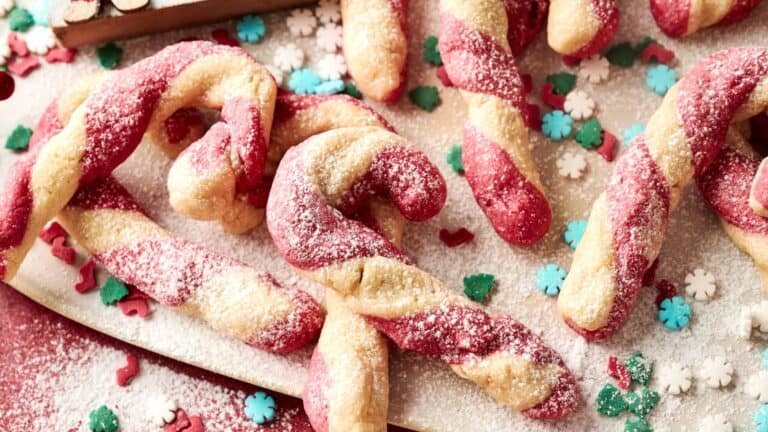 A close-up of red and white twisted candy cane-shaped cookies dusted with powdered sugar, surrounded by festive sprinkles and sugar decorations.