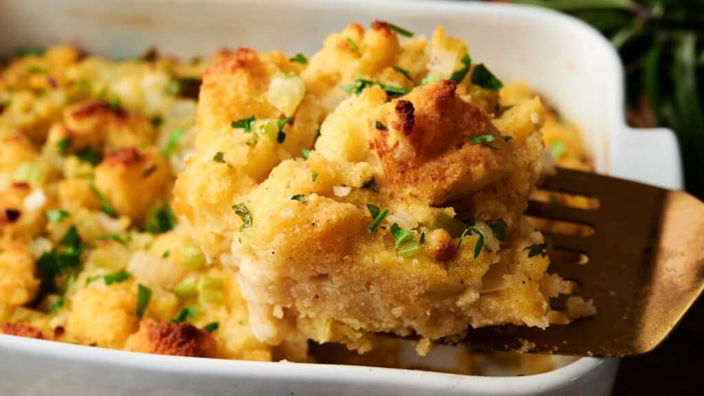 A serving of baked bread stuffing with herbs and vegetables is lifted from a white casserole dish with a spatula.