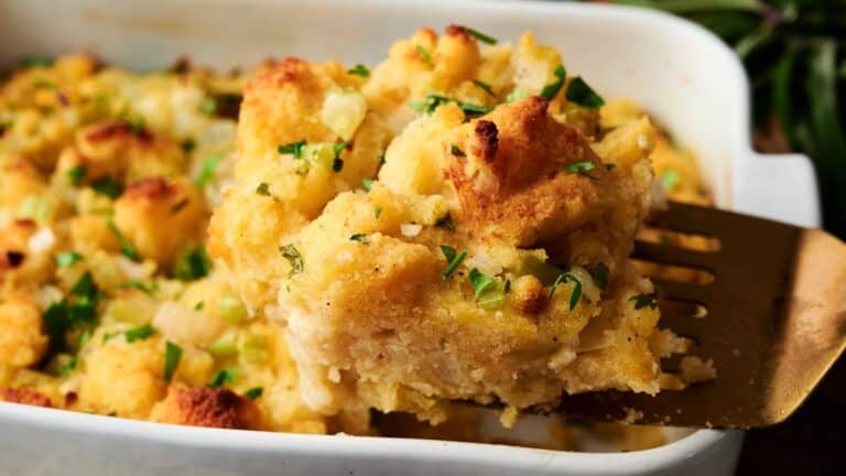 A serving of baked bread stuffing with herbs and vegetables is lifted from a white casserole dish with a spatula.