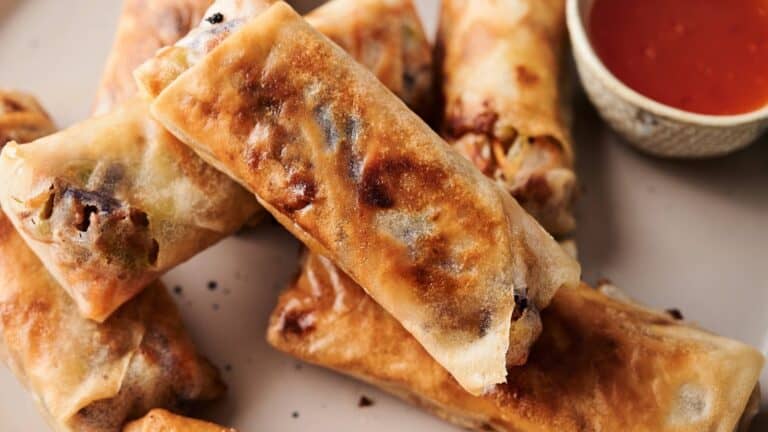 A close-up of several golden-brown spring rolls on a plate, with a small bowl of red dipping sauce on the side.