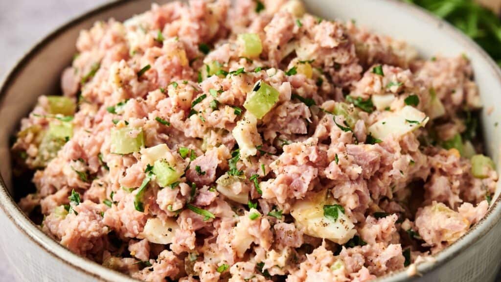 A bowl filled with ham salad mixed with chopped celery, hard-boiled eggs, herbs, and a creamy dressing.