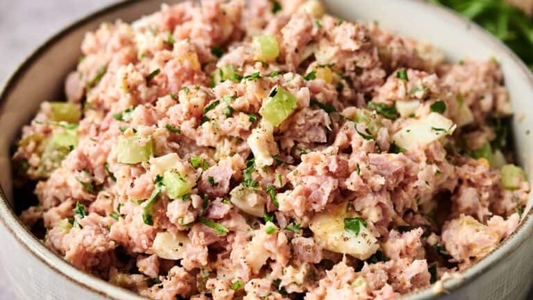 A bowl filled with ham salad mixed with chopped celery, hard-boiled eggs, herbs, and a creamy dressing.