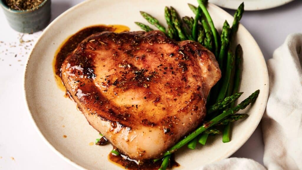 A cooked pork chop with a brown glaze served on a plate with steamed asparagus spears, garnished with ground black pepper.