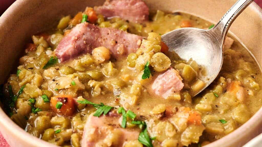 A bowl of split pea soup with chunks of ham, diced carrots, and parsley, with a spoon resting inside.