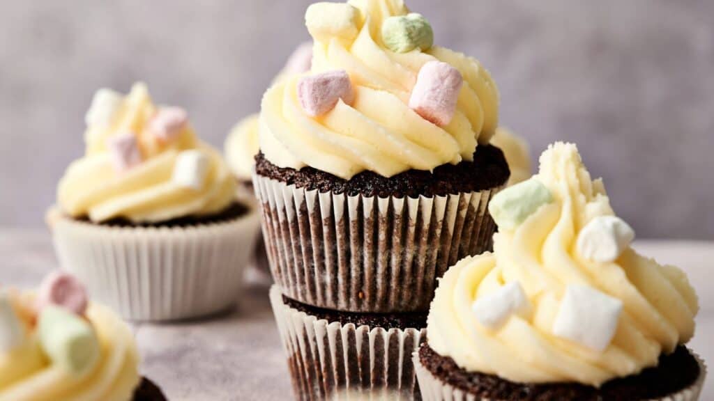 Chocolate cupcakes with white frosting are topped with small pastel-colored marshmallows and arranged on a light surface.