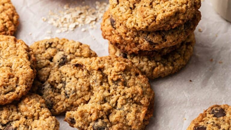 A stack of oatmeal chocolate chip cookies sits on parchment paper, with several cookies scattered around, one showing a bite taken out.