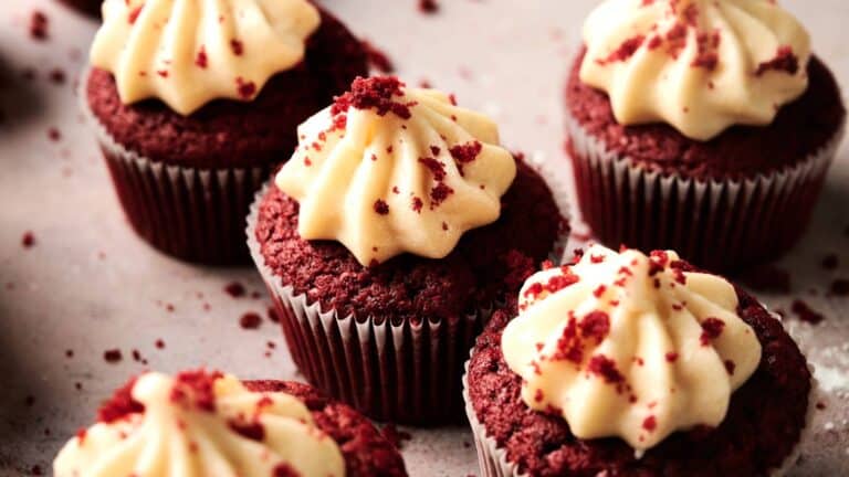 Red velvet cupcakes with cream cheese frosting and red crumbs on top are arranged on a light surface.