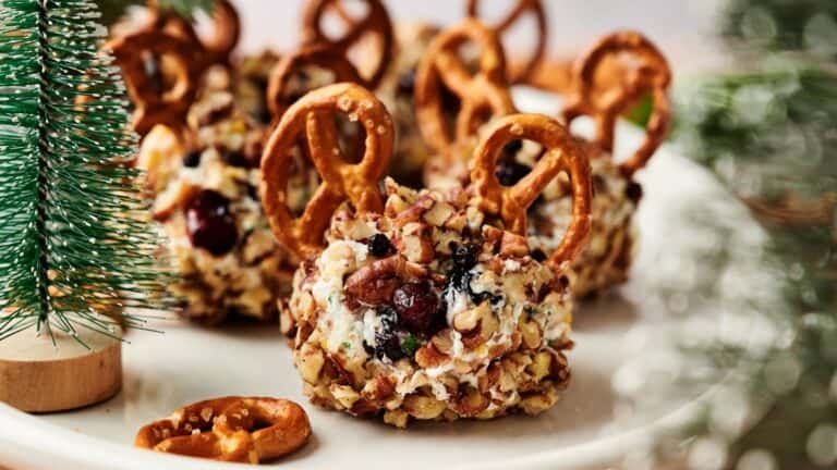 Round cheese balls coated with chopped nuts and dried fruit, each decorated with pretzel twists, arranged on a white plate near a small artificial pine tree.