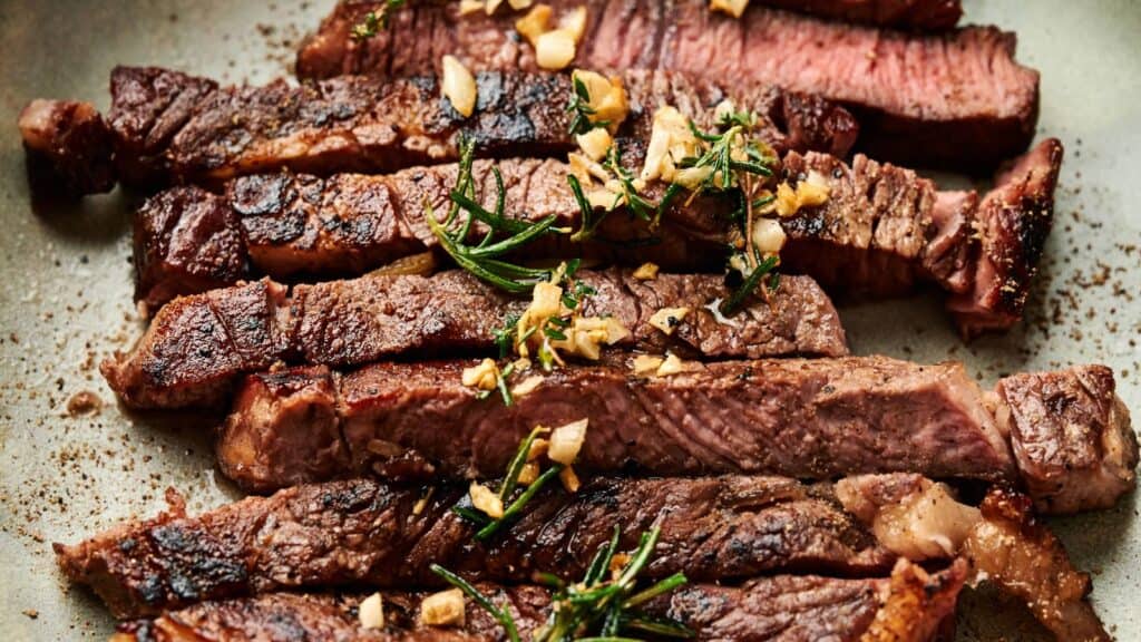 Sliced grilled steak topped with chopped garlic and sprigs of rosemary on a plate.