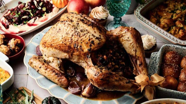 Roast turkey filled with stuffing is served on a platter, surrounded by various side dishes, bread rolls, and desserts on a dining table.