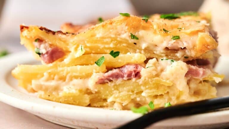 A close-up of a slice of potato gratin with layers of potatoes, ham, creamy sauce, and melted cheese, garnished with chopped parsley on a white plate.