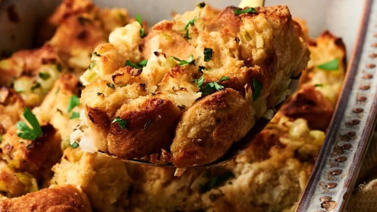 A serving spoon holds a piece of golden-brown bread stuffing garnished with chopped herbs, with more stuffing visible in the background.