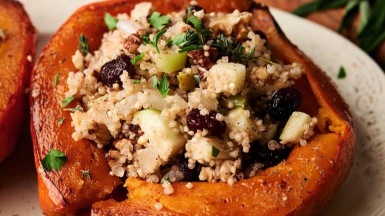 Roasted acorn squash half filled with a stuffing of couscous, chopped apples, dried cranberries, walnuts, and fresh herbs, served on a white plate.
