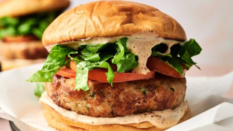 A close-up of a chicken burger with lettuce, tomato, and creamy sauce in a toasted bun, served on white paper.