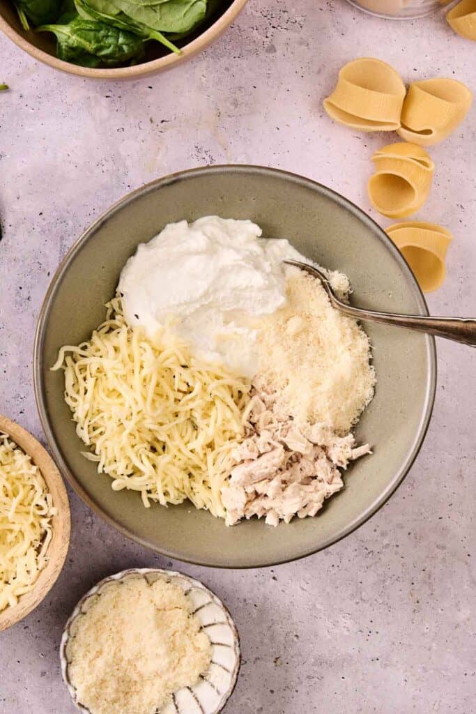 A bowl contains ingredients for Marry Me Chicken Stuffed Shells: shredded mozzarella, grated Parmesan, shredded cooked chicken, and a dollop of ricotta cheese with a spoon. Uncooked pasta shells and spinach are nearby.