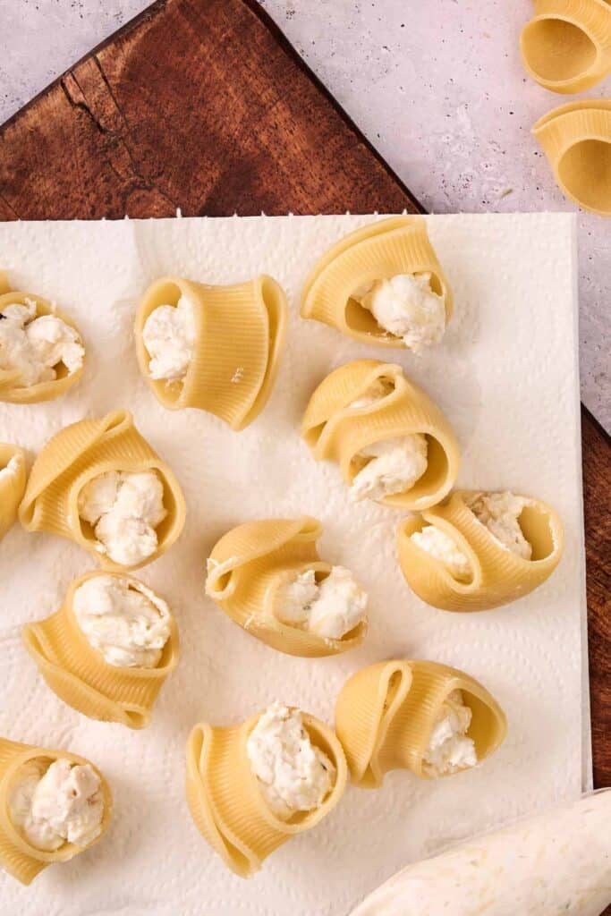 Uncooked pasta shells filled with a creamy Marry Me Chicken Stuffed Shells ricotta mixture are arranged on a paper towel, with a wooden board visible underneath.