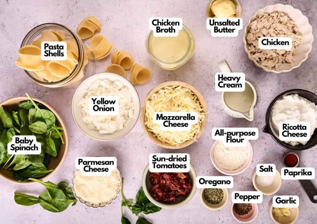 Top-down view of labeled ingredients for Marry Me Chicken Stuffed Shells in bowls and plates, including chicken, pasta shells, cheeses, spinach, sun-dried tomatoes, spices, and more on a light surface.