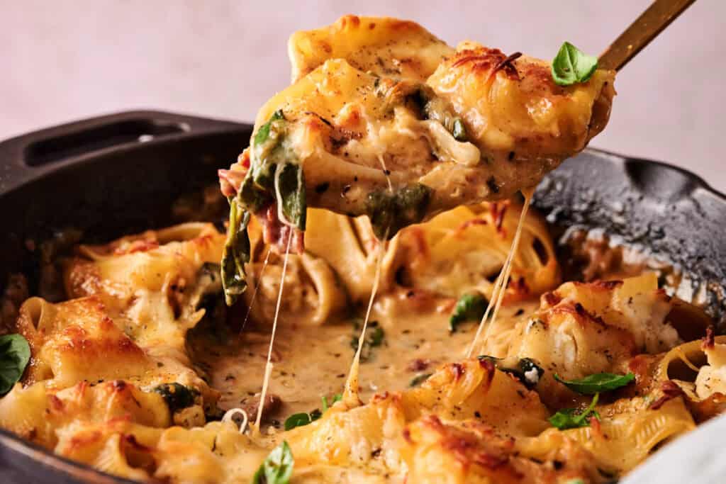 A serving spoon lifts Marry Me Chicken Stuffed Shells with spinach and sauce from a cast iron skillet, with melted cheese stretching.
