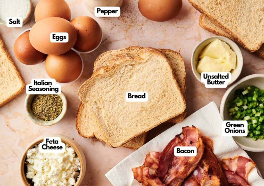 Various ingredients for Sheet Pan Eggs&mdash;including eggs, bread slices, bacon, feta cheese, green onions, unsalted butter, Italian seasoning, salt, and pepper&mdash;are laid out on a countertop.