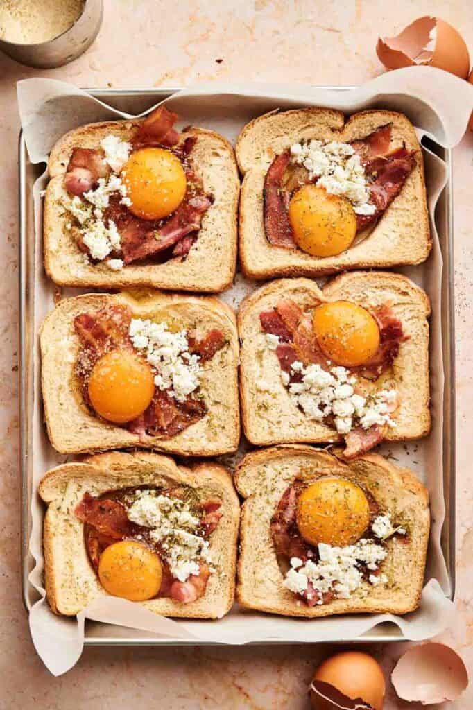 Six slices of bread on a baking tray, each topped with bacon, a raw egg yolk, and crumbled cheese&mdash;perfect for making easy Sheet Pan Eggs. Eggshells are nearby on the countertop, ready for cleanup.