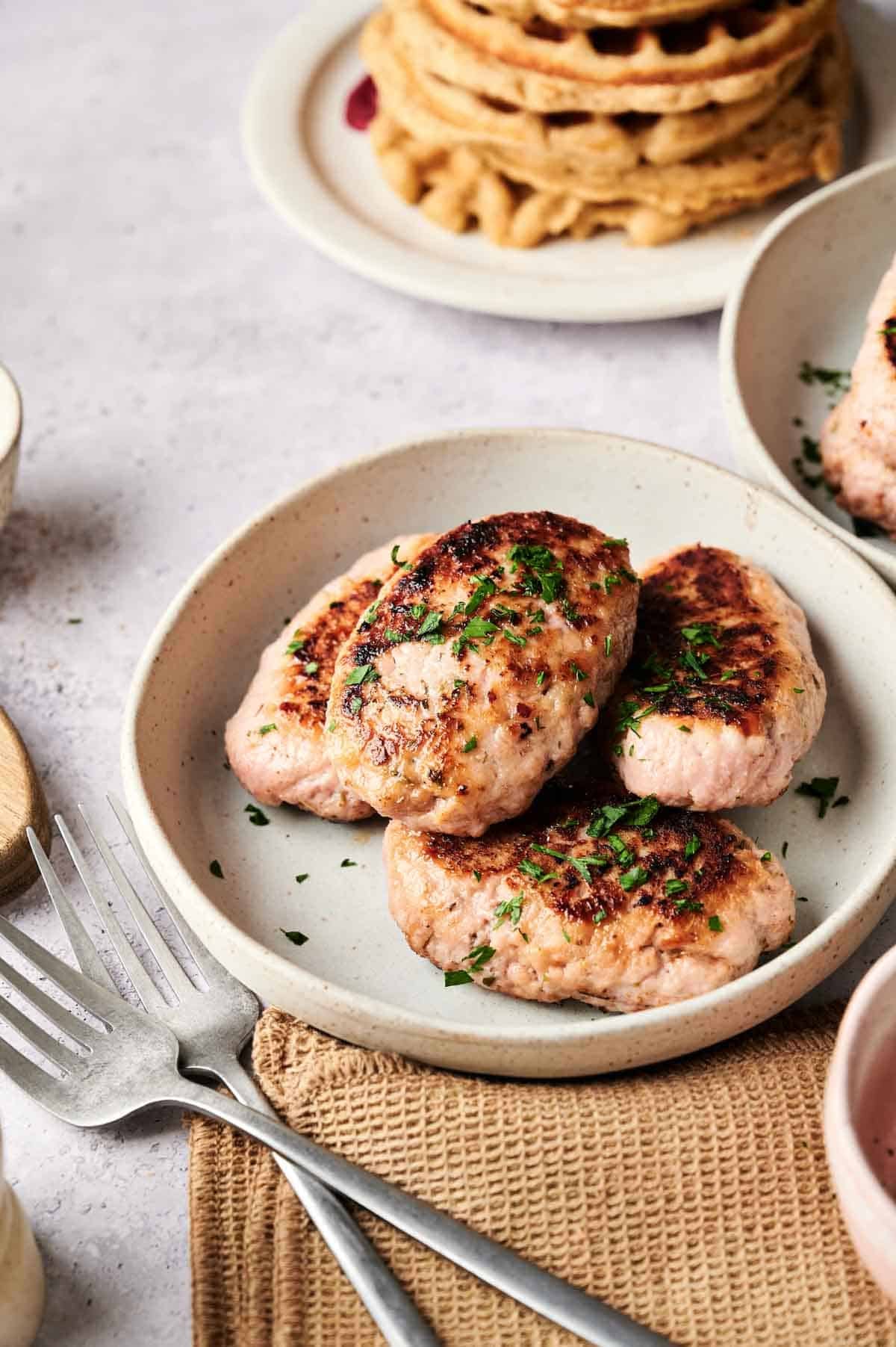 A plate with three browned turkey sausage patties garnished with chopped herbs sits next to two forks, with waffles visible in the background.