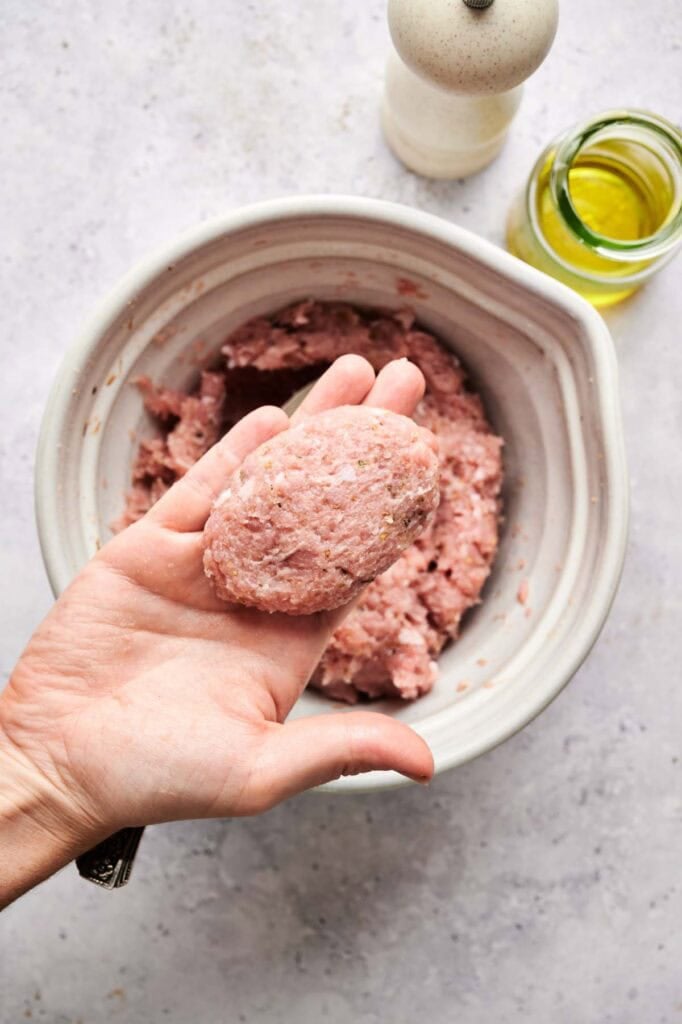 A hand holds an oval-shaped portion of raw ground meat mixture over a bowl of more meat, ready to form Turkey Sausage Patties, with a pepper grinder and a jar of oil nearby.