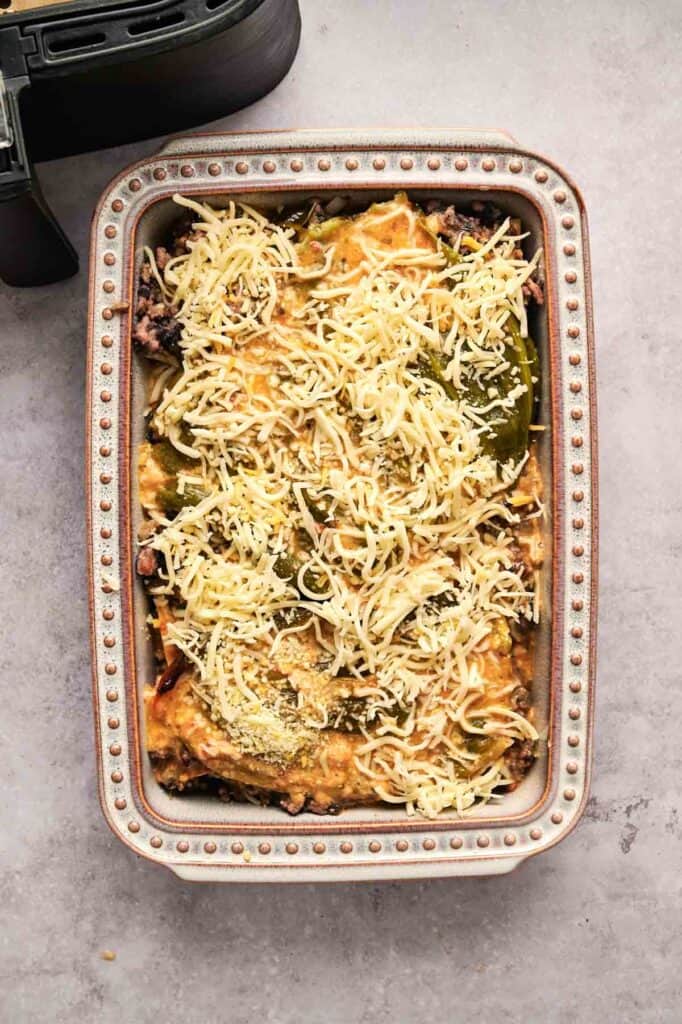 A rectangular baking dish filled with layered lasagna, topped with shredded cheese, sits on a gray countertop next to part of a black Air Fryer Chili Relleno Casserole appliance.