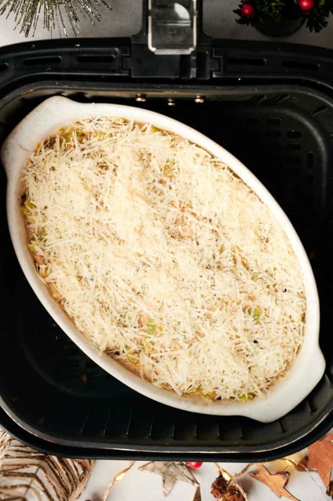 A white oval baking dish filled with Air Fryer Leek Christmas Casserole, topped with shredded cheese, sits inside an air fryer basket, ready to be cooked.