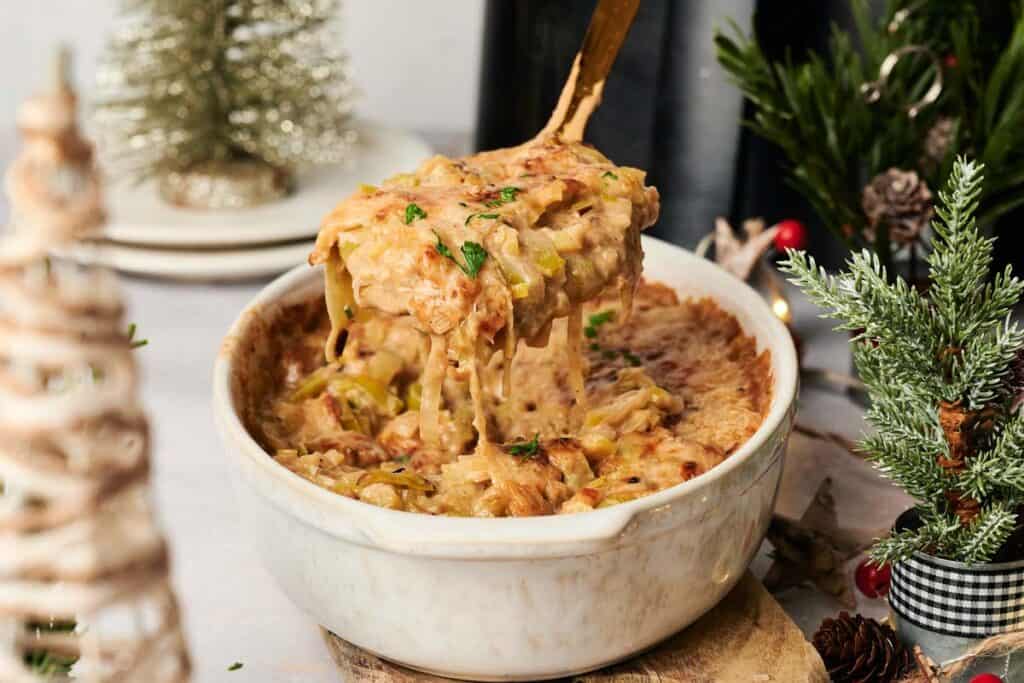 A serving spoon lifts a portion of cheesy Air Fryer Leek Christmas Casserole from a white dish, with holiday decorations in the background.