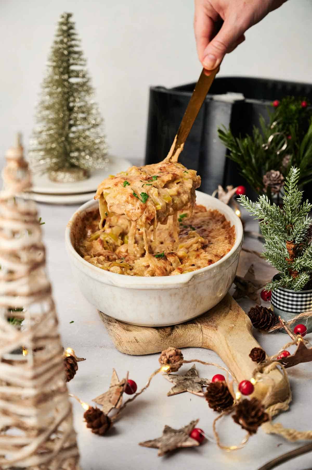 A hand lifts a serving of Air Fryer Leek Christmas Casserole from a white bowl, surrounded by small Christmas trees and festive holiday decorations.