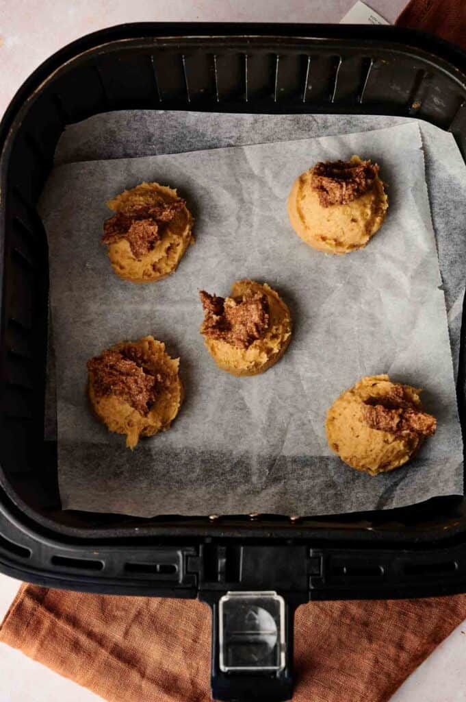 Five scoops of cookie dough, some topped with a brown mixture, placed on parchment paper in an air fryer basket—perfect for making Air Fryer Crumbl Cinnamon Swirl Cookies.